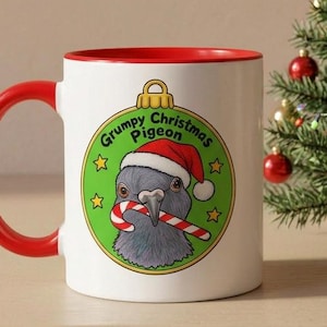 Könnte beinhalten: Ein weißer Keramikbecher mit rotem Henkel und Rand, verziert mit einer Grafik eines grauen Vogels mit Weihnachtsmütze, der eine Zuckerstange in einer grünen Kugel hält, dazu der Text Grumpy Christmas Pigeon und kleine gelbe Sterne.