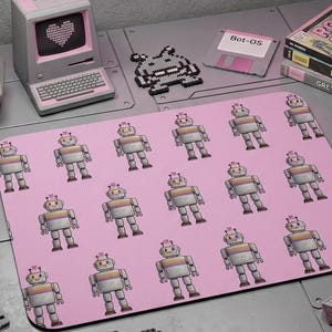 Könnte beinhalten: Ein rechteckiges Mauspad mit einem wiederkehrenden Muster aus kleinen grauen und orangefarbenen Robotern auf hellrosa Hintergrund. Das Schreibtisch-Setup umfasst ein rosa Neonschild mit der Aufschrift CTRL+ALT+NERD, einen Mini-Computer, Disketten und eine Roboterfigur.