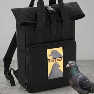 Könnte beinhalten: Ein schwarzer Rolltop-Rucksack mit zwei Tragegriffen und einem schwarzen Kunststoff-Schnallenverschluss. Die Vordertasche zeigt eine rechteckige Grafik mit zwei Tauben-Illustrationen und dem Text PIGEON in roten Buchstaben auf gelbem Grund.