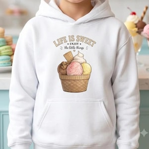 Op de afbeelding: Een witte hoodie met een afbeelding van een ijshoorntje met drie bollen in bruin, roze en geel, bekroond met een wafel. De tekst boven de afbeelding luidt LIFE IS SWEET ENJOY the little things in een bruin, gebogen lettertype.