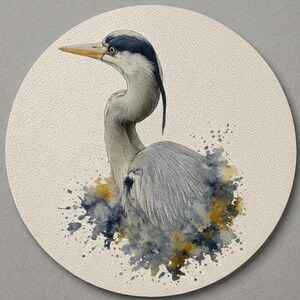 May include: A circular coaster featuring a watercolor illustration of a grey heron. The bird has a long yellow beak, a black crest, and grey plumage. The base of the heron is surrounded by abstract splashes of dark blue, grey, and ochre paint.
