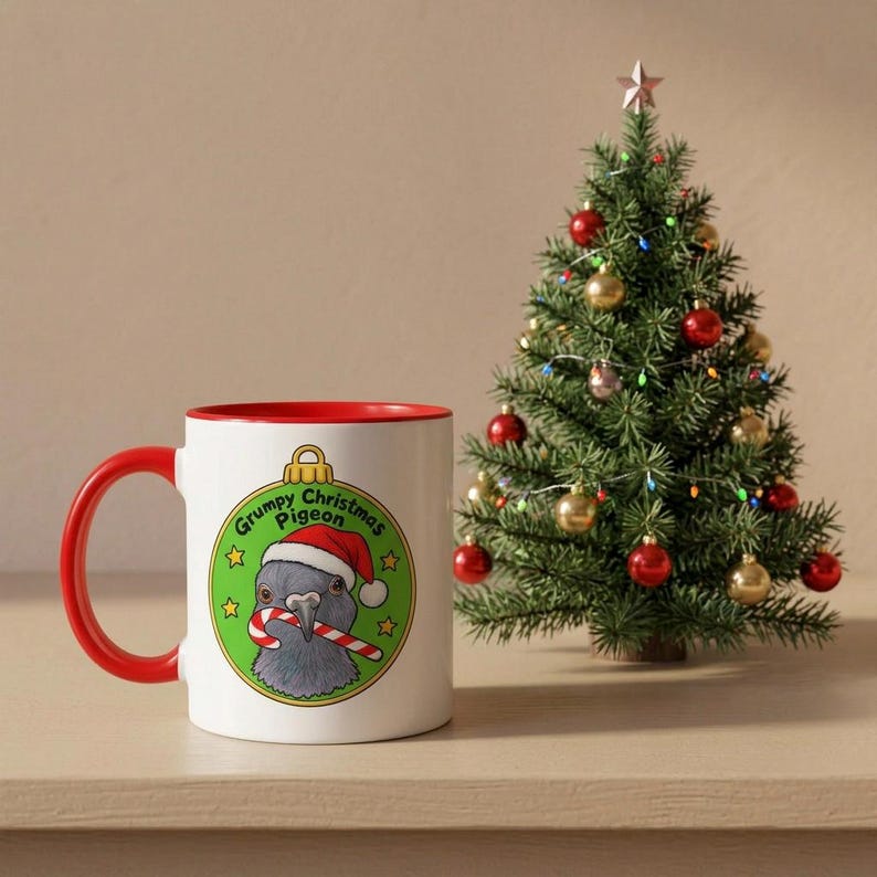 May include: A white ceramic mug with a red handle and rim, featuring a graphic of a grey pigeon wearing a Santa hat and holding a candy cane inside a green ornament shape with the text Grumpy Christmas Pigeon and small yellow stars.