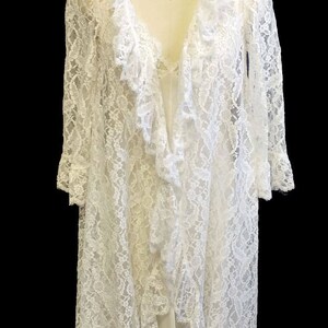 K&ouml;nnte beinhalten: Ein wei&szlig;es Spitzenkleid mit R&uuml;schenkragen und langen &Auml;rmeln. Das Kleid ist vorne offen und zeigt darunter ein wei&szlig;es Unterkleid. Das Kleid befindet sich auf einer Puppe mit einer Strasskette.