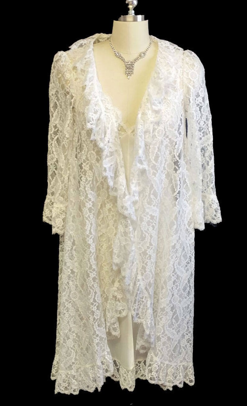 K&ouml;nnte beinhalten: Ein wei&szlig;es Spitzenkleid mit R&uuml;schenkragen und langen &Auml;rmeln. Das Kleid ist vorne offen und zeigt darunter ein wei&szlig;es Unterkleid. Das Kleid befindet sich auf einer Puppe mit einer Strasskette.