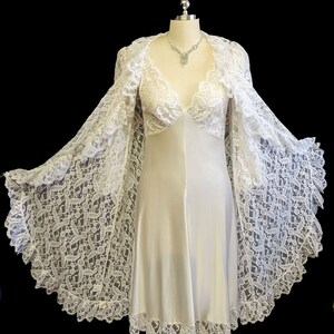 K&ouml;nnte beinhalten: Ein wei&szlig;es Satin-Schlafkleid mit einer Spitzen&uuml;berlage und einem passenden Spitzen-Morgenmantel. Das Kleid hat einen V-Ausschnitt und ein tailliertes Oberteil. Der Morgenmantel hat lange &Auml;rmel und eine flie&szlig;ende Silhouette.