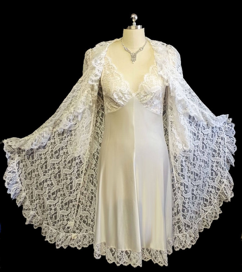 K&ouml;nnte beinhalten: Ein wei&szlig;es Satin-Schlafkleid mit einer Spitzen&uuml;berlage und einem passenden Spitzen-Morgenmantel. Das Kleid hat einen V-Ausschnitt und ein tailliertes Oberteil. Der Morgenmantel hat lange &Auml;rmel und eine flie&szlig;ende Silhouette.