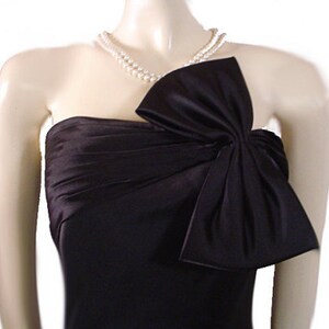 Op de afbeelding: Een zwarte strapless jurk met een groot strikdetail op het lijfje. De jurk is gemaakt van een glanzende stof en is geaccessoireerd met een parelketting met dubbele streng. De jurk wordt op een mannequin getoond.