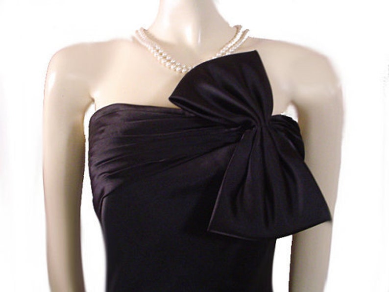 Op de afbeelding: Een zwarte strapless jurk met een groot strikdetail op het lijfje. De jurk is gemaakt van een glanzende stof en is geaccessoireerd met een parelketting met dubbele streng. De jurk wordt op een mannequin getoond.