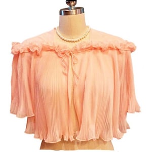 May include: A peach-colored, pleated cape-style top with a ruffled neckline and short sleeves. The top is displayed on a mannequin and features a delicate tie closure at the chest. A pearl necklace is also visible.