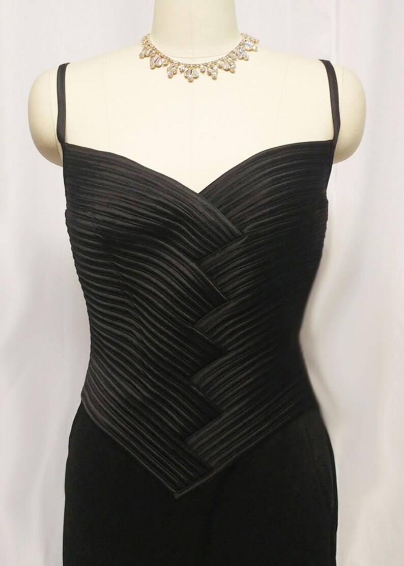 May include: A black satin evening gown with a pleated bodice and a sweetheart neckline. The gown has thin spaghetti straps and a fitted silhouette.