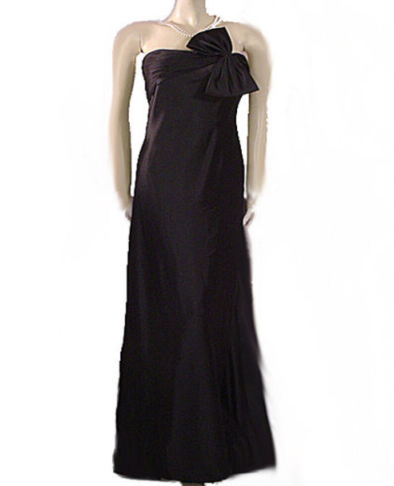 Op de afbeelding: Een zwarte, strapless avondjurk, vloerlang. De jurk heeft een grote strik op het lijfje en een zwierige rok. Een parelketting is over de schouder gedrapeerd. De jurk wordt op een mannequin getoond.