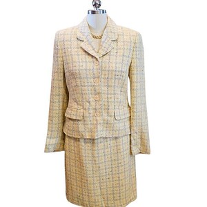 May include: A two-piece skirt suit in a light yellow tweed fabric with a subtle plaid pattern. The jacket has a notched collar, two front pockets, and button closures. The skirt is knee-length. A gold-colored necklace is visible.
