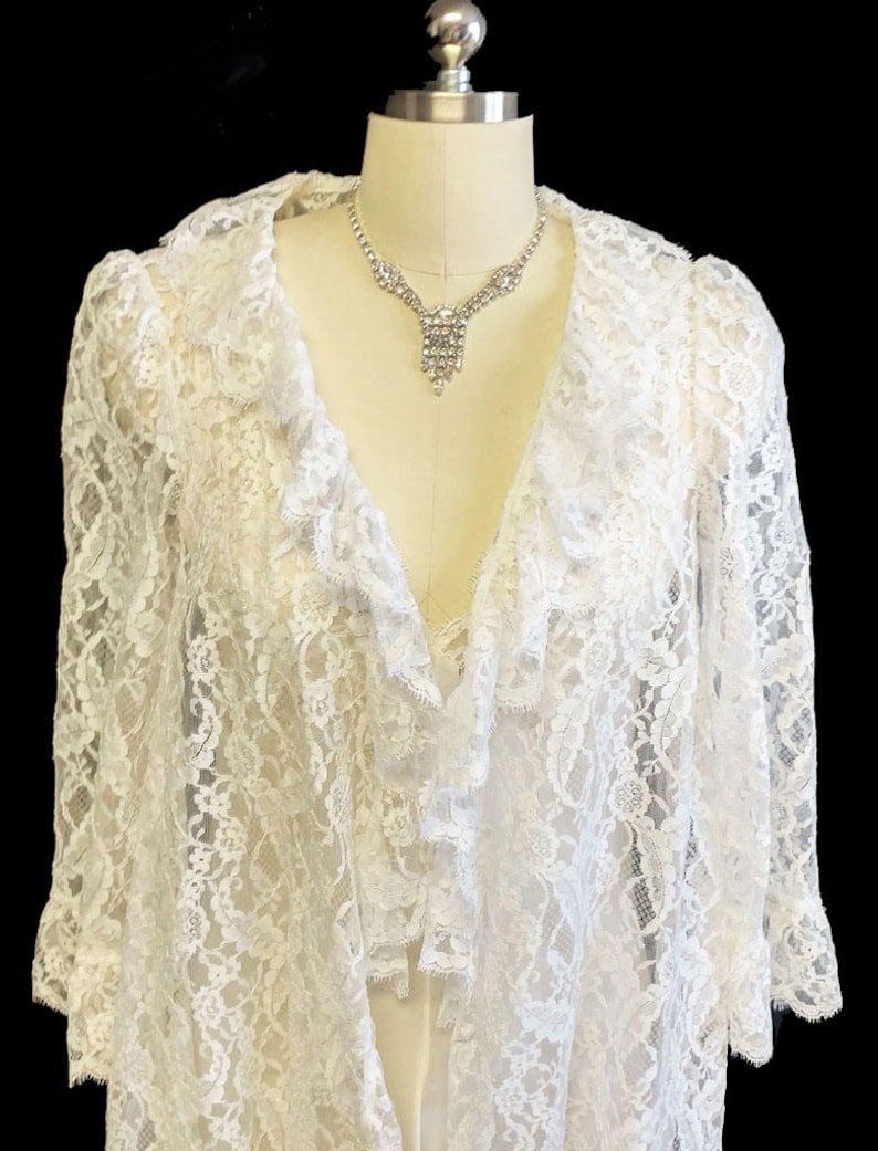 K&ouml;nnte beinhalten: Wei&szlig;e Spitzenrobe mit R&uuml;schenkragen und langen &Auml;rmeln. Die Robe befindet sich auf einem Mannequin und hat eine Strasskette.