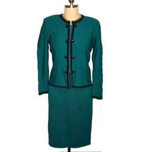 May include: Teal knit two-piece suit. The jacket has long sleeves, a round neckline, and black trim. The skirt is knee-length. The jacket features black buttons and trim. The suit is displayed on a mannequin.