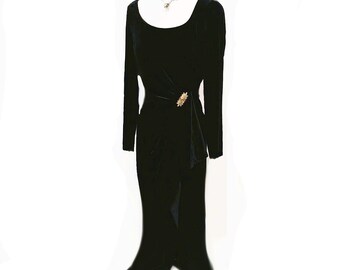 May include: A long black velvet dress with long sleeves and a scoop neckline. The dress features a draped waist detail and a gold-colored brooch. The skirt flares out at the bottom. A silver necklace is visible at the neckline.