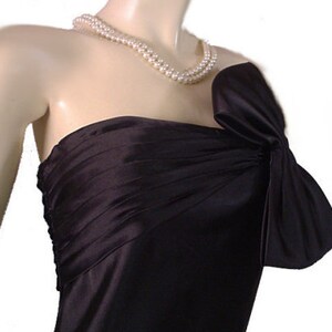 Op de afbeelding: Een zwarte strapless jurk met een strikdetail op de schouder. De jurk is gemaakt van een glanzende stof en wordt gecombineerd met een parelketting met dubbele streng. De jurk wordt getoond op een mannequin.