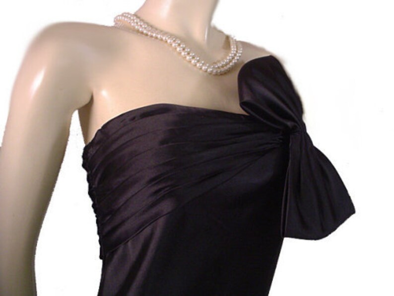 Op de afbeelding: Een zwarte strapless jurk met een strikdetail op de schouder. De jurk is gemaakt van een glanzende stof en wordt gecombineerd met een parelketting met dubbele streng. De jurk wordt getoond op een mannequin.