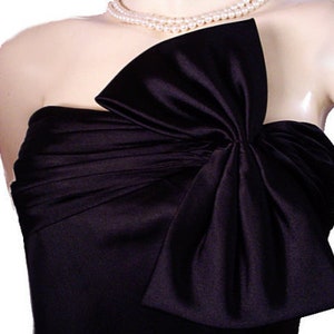 Op de afbeelding: Zwarte strapless satijnen jurk met een grote strik op het lijfje. De jurk wordt gedragen met een parelketting. De stof heeft een lichte glans en de strik voegt een dramatisch element toe aan de formele kleding.