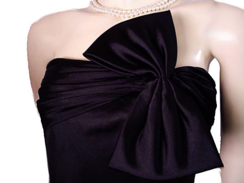 Op de afbeelding: Zwarte strapless satijnen jurk met een grote strik op het lijfje. De jurk wordt gedragen met een parelketting. De stof heeft een lichte glans en de strik voegt een dramatisch element toe aan de formele kleding.