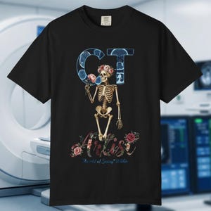 Op de afbeelding: Een zwart T-shirt met korte mouwen en een afbeelding van een skelet met een bloemenkrans dat een roze roos vasthoudt. De letters CT zijn gevuld met medische scanbeelden. Daaronder staat het woord Artist in bloemrijk schrift met de tekst The Art of Seeing Within.