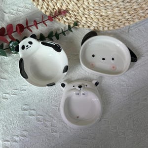 Puede incluir: Tres platos pequeños de cerámica con forma de caras de animales. Un plato presenta un diseño de panda en blanco y negro, otro es una cara de perro blanca con orejas negras y el tercero es una cara de oso blanca con patas en el borde.