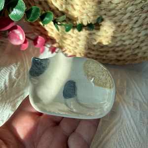 Può includere: Un piccolo piattino in ceramica bianca a forma di gatto raggomitolato. Il piattino presenta dettagli dipinti a mano, tra cui una macchia grigia sulla testa, una coda grigia e una macchia beige testurizzata sul dorso, con finitura lucida.
