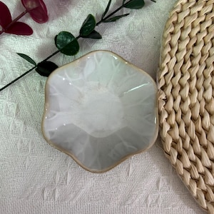 May include: A small, hexagonal ceramic dish with a glossy white glaze and a light brown rim. The dish features a subtle, wavy, petal-like edge design. The surface of the ceramic has a smooth, reflective finish that catches the light, highlighting the clean, geometric shape of the bowl.