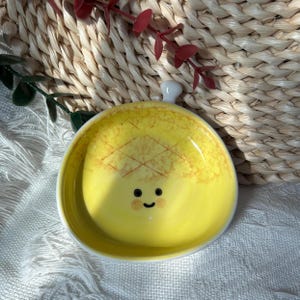 Peut inclure: Un petit plat rond en céramique jaune orné d'un motif d'ananas peint à la main à l'intérieur. Le plat présente un visage souriant simple avec deux yeux en points noirs et une bouche courbée, ainsi qu'une petite protubérance blanche sur le bord.