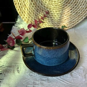May include: A ceramic tea cup and saucer set featuring a deep blue speckled glaze with dark brown rim accents. The cup has a sturdy rectangular handle and a smooth, rounded body, while the matching saucer has a wide, flat rim design.
