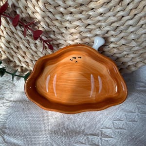 May include: A ceramic dish shaped like a piece of cooked meat with a bone handle. The dish features a glossy orange finish with painted white highlights and a small, simple black facial expression consisting of two eyes and a mouth.