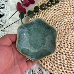 Könnte beinhalten: Eine kleine, blumenförmige Keramikschale mit einer reaktiven Glasur in gesprenkelten Schattierungen von tiefem Waldgrün, Petrol und helleren Salbei-Akzenten. Die Schale hat eine glatte, glänzende Oberfläche mit einem leicht unregelmäßigen Rand.