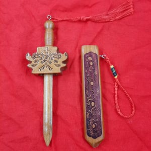 Peut inclure: Une épée en bois avec son fourreau assorti. La garde de l'épée est gravée d'un symbole yin-yang et de trigrammes, et est attachée à un pompon rouge. Le fourreau présente une gravure de dragon complexe et une boucle en cordon rouge.