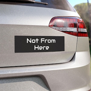May include: A rectangular black bumper sticker with white text that reads Not From Here, applied to the silver boot of a vehicle. The sticker features a clean, sans-serif font and is positioned horizontally below the rear window and taillight.