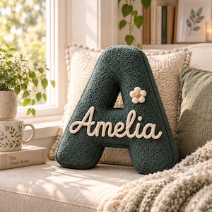 May include: A dark green, textured, letter-shaped throw cushion in the form of the letter A. The cushion features the name Amelia in white cursive script lettering across the front and a small, cream-coloured, six-petaled flower accent.