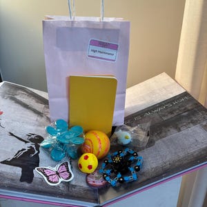Könnte beinhalten: Eine hellrosa Geschenktüte aus Papier mit einem weißen Namensschild-Aufkleber mit der Aufschrift Hello High Maintenance, einem gelben Notizbuch, einer blauen Blumen-Haarklammer, einem gelb-orange gestreiften Ball, einem kleinen gelben Ball und einem schwarzen Fidget Spinner.