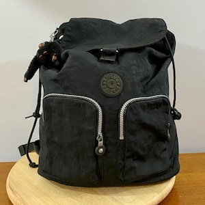 May include: A black nylon backpack featuring a top flap closure, two front zippered pockets with silver-toned hardware, and a circular logo patch centered on the front. A small, fuzzy black monkey keychain is attached to the side strap.