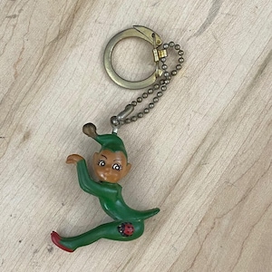 Puede incluir: Un llavero con una pequeña figura de elfo vestida de verde, con cara bronceada, sombrero puntiagudo y zapatos con puntas rojas. El elfo está en posición sentada con un detalle de mariquita en la pierna, unido a una cadena y una anilla.