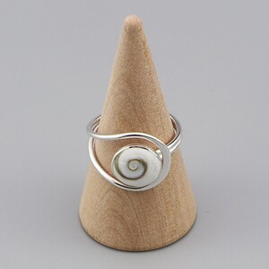 Puede incluir: Un anillo de plata con una incrustación de concha en espiral de color blanco y gris. La banda está compuesta por dos hilos de plata finos y lisos que se curvan alrededor del elemento central, creando un diseño abierto y asimétrico.