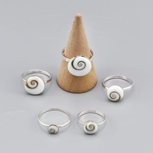 Puede incluir: Cinco anillos de plata con incrustaciones circulares de concha de ojo de Shiva con un patrón de espiral natural. Un anillo se muestra en un soporte cónico beige, mientras que los otros cuatro anillos están dispuestos alrededor sobre una superficie gris.