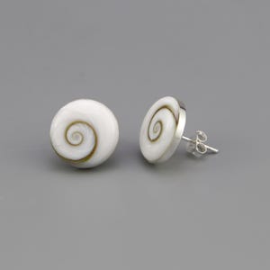 Puede incluir: Un par de pendientes de botón con conchas de ojo de shiva circulares que presentan un patrón de espiral natural en blanco y marrón. Cada concha está engastada en una montura de metal plateado con un cierre de poste estándar para la oreja.