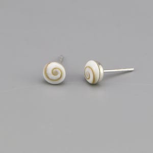 May include: A pair of stud earrings featuring circular shiva shell cabochons with a natural white and tan spiral pattern. Each shell is set in a polished silver-tone metal base with a straight post backing for secure attachment to the earlobe.