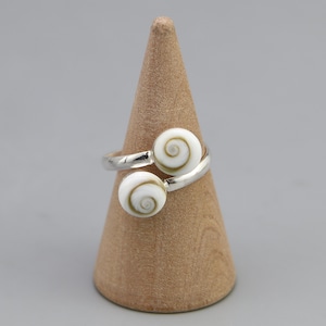 Puede incluir: Un anillo abierto con dos cabujones redondos de concha de ojo de Shiva blanca, que presentan un patrón de espiral marrón natural. La banda del anillo está hecha de metal pulido en tono plateado, creando un acabado liso y brillante.