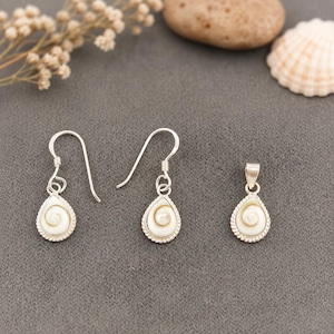 Könnte beinhalten: Ein Schmuckset bestehend aus zwei Ohrhängern und einem Anhänger, jeweils mit einer tropfenförmigen Shiva-Muschel mit natürlichem Spiralmuster, eingefasst in eine silberfarbene Metallfassung mit gedrehtem Rand und silbernen Haken.