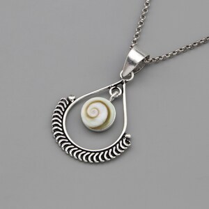 May include: A silver-toned pendant necklace featuring a teardrop-shaped frame with a textured, scalloped edge. Suspended within the centre of the frame is a small, circular shiva eye shell with a natural white and brown spiral pattern.