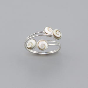 Puede incluir: Un anillo abierto de tono plateado con cuatro pequeños acentos de conchas circulares en espiral de color blanco y marrón. La banda está formada por dos hilos metálicos finos y paralelos que se curvan para sostener las conchas en su lugar.
