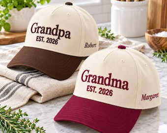 Könnte beinhalten: Zwei Baseballkappen mit cremefarbenen Kronen und farbigen Schirmen. Eine Kappe hat einen braunen Schirm mit brauner Stickerei, die Grandpa EST. 2026 und Robert an der Seite zeigt. Die andere hat einen weinroten Schirm mit weinroter Stickerei, die Grandma EST. 2026 und Margaret an der Seite zeigt.