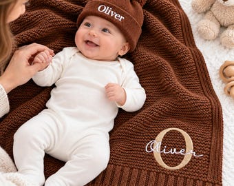 Könnte beinhalten: Ein Baby trägt einen weißen Langarm-Body und eine braune Strickmütze mit Bommel, auf der der Name Oliver in Weiß eingestickt ist. Das Baby liegt auf einer braunen Strickdecke, die mit dem Namen Oliver und einem großen Buchstaben O bestickt ist.