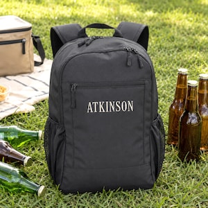 May include: A black backpack featuring the word ATKINSON printed in white on the front pocket. The backpack has a top handle, two shoulder straps, and side mesh pockets. It is constructed with multiple zippered compartments and durable fabric.