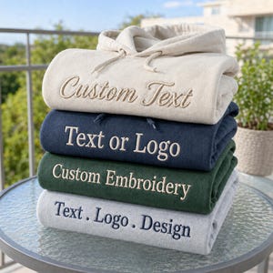 May include: A stack of four folded hooded sweatshirts in cream, navy blue, dark green, and light gray. Each hoodie features centered embroidery: the cream one reads Custom Text, the navy reads Text or Logo, the green reads Custom Embroidery, and the gray reads Text . Logo . Design.