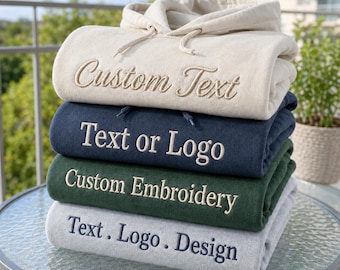 Könnte beinhalten: Ein Stapel aus vier gefalteten Kapuzenpullovern in Creme, Marineblau, Dunkelgrün und Hellgrau. Jeder Hoodie hat eine mittige Stickerei: der cremefarbene zeigt Custom Text, der marineblaue Text or Logo, der grüne Custom Embroidery und der graue Text . Logo . Design.